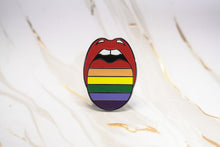 Load image into Gallery viewer, A tongue-shaped enamel pin featuring horizontal rainbow pride stripes with red lips and white teeth, displayed on a white marble surface with gold veining.
